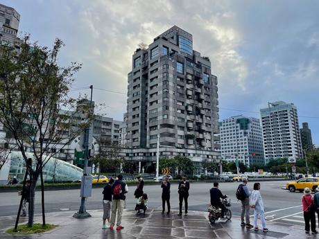 A bustling city street with a prominent building in the center featuring a unique staggered architectural design, pedestrians and vehicles in the foreground.