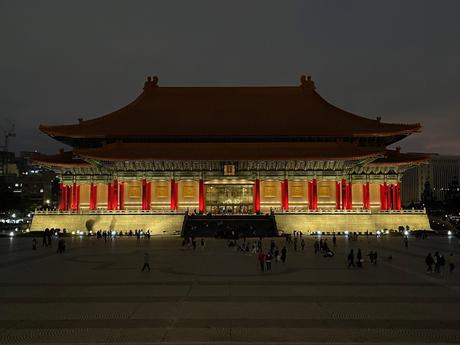 The National Concert Hall in Taipei lit up at night, with bright red columns and golden tones reflecting against the dark sky, and visitors walking around the plaza.