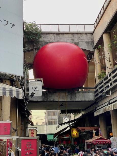 A large, bright red inflatable ball art installation wedged between two buildings in a busy outdoor market. Crowds of people are shopping and walking below it.