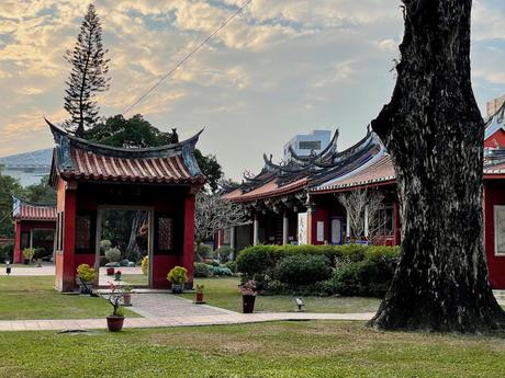 The serene courtyard of the Tainan Confuscious Temple, with traditional red buildings with ornate roofs, surrounded by green grass and trees under a partly cloudy evening sky.