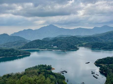 A tranquil lake surrounded by forested hills and mountains under a cloudy sky, with scattered boats visible on the water's surface.