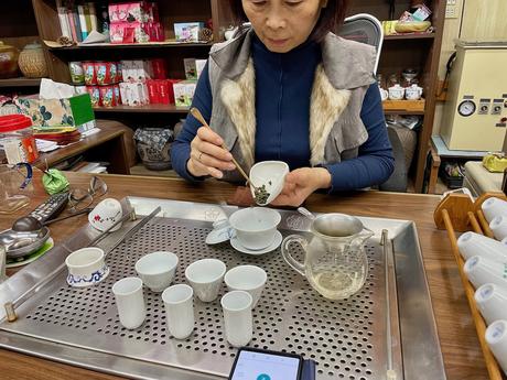 A woman preparing oolong tea at a wooden table, carefully handling tea leaves and surrounded by traditional cups, teapots, and brewing equipment.