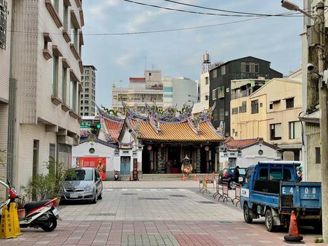 A wide view of a Taiwanese temple at the end of a street, surrounded by urban buildings, with a clear view of its intricately decorated roof and gates.