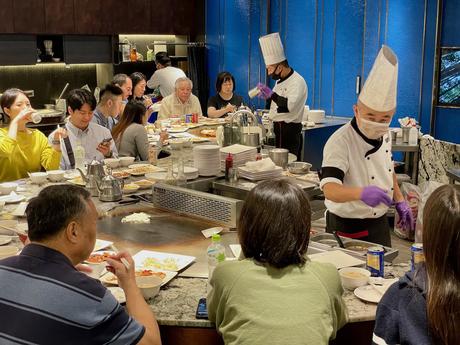 Diners seated around a teppanyaki table while chefs in tall hats perform live cooking, with sizzling dishes and a lively atmosphere.