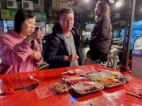 A man and a woman seated at a red table outdoors, enjoying trays of colorful sashimi, surrounded by a lively night market environment.