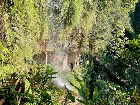 A green, mossy cliffside with a waterfall spraying mist over the surrounding foliage, and a stone pathway leading to an old temple built into the rocks.