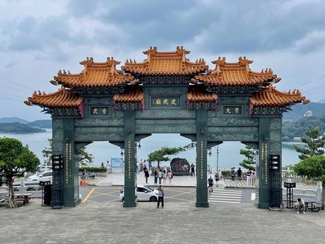 A large traditional Chinese-style gate at Wenwu Temple, Taiwan, with ornate orange roof tiles and green pillars, overlooking a lake and a scenic view of mountains in the distance.