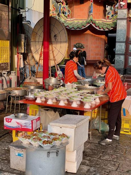 Two women preparing traditional Taiwanese dishes at an outdoor food stall near a temple. Fresh plates of greens and meats are laid out on a table in the golden evening light.