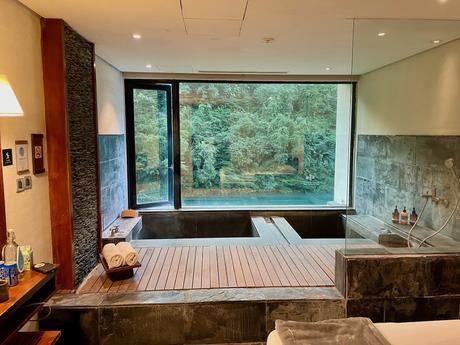 A luxurious bathroom with a stone and wood design, featuring a sunken bathtub by a large window offering a view of lush green trees outside.