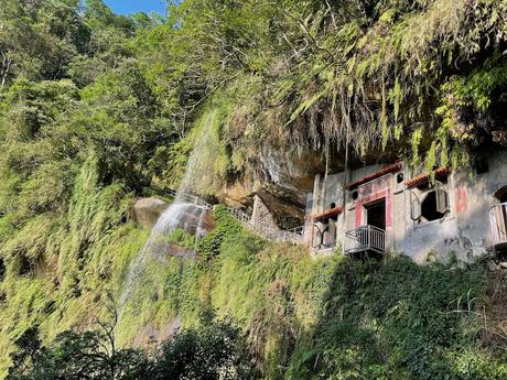 A picturesque view of a waterfall cascading over a lush green cliff, next to an old stone temple with red doors and balconies.