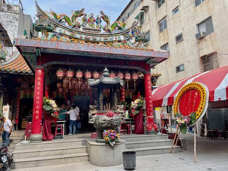 A Taiwanese temple decorated with intricate carvings, colorful lanterns, and dragon sculptures on the rooftop. A circular floral wreath with red and yellow accents stands beside it.