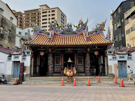 A traditional Taiwanese temple with a detailed, colorful rooftop featuring statues of dragons and deities, framed by modern apartment buildings in the background.