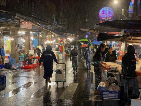 A bustling seafood market at night with vendors selling fresh fish and shoppers browsing under umbrellas and market awnings, while rain reflects off the wet ground.