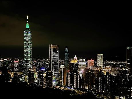 A stunning nightscape featuring Taipei 101 illuminated in green, surrounded by other city buildings glowing with lights under a dark sky.