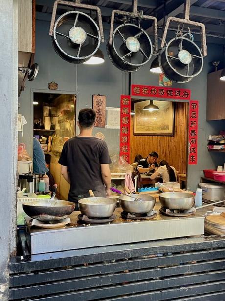 A street food vendor's stall with large woks on display, a man cooking in the foreground, and a cozy dining area with patrons in the background.