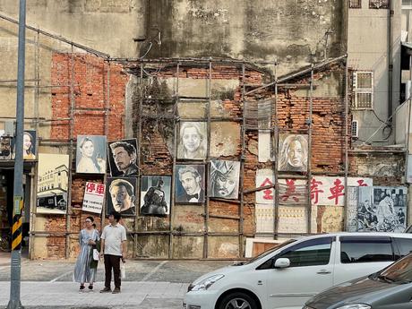 A rustic brick wall adorned with paintings of iconic movie characters, including portraits of Johnny Depp and Robert Downey Jr. Two people stand nearby in front of parked cars.