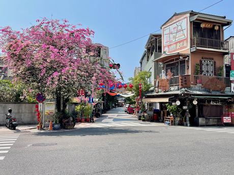 A vibrant street corner in Tainan, featuring a blooming pink bougainvillea tree, traditional buildings, and colorful decorations hanging across the street.