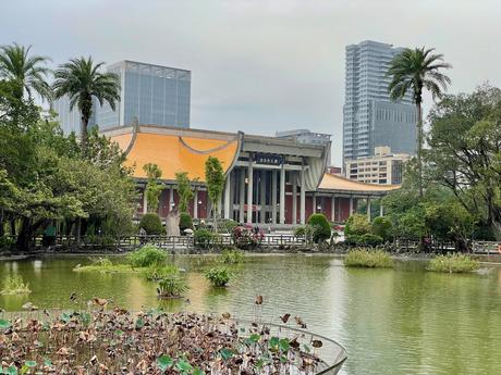 A pond with lotus plants in front of a grand traditional building with an orange roof, surrounded by palm trees and modern skyscrapers in the background.