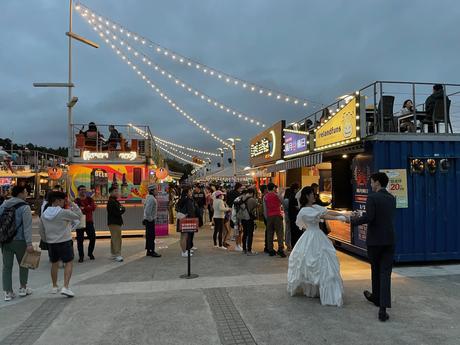 A lively outdoor food market at night with string lights illuminating various colorful food stalls, a bride in a white wedding dress and a groom in a suit.