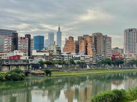 A scenic view of a city skyline with Taipei 101 in the background, framed by a calm river reflecting the buildings under a cloudy sky.