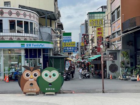 A typical street in Taiwan lined with shops and signs in various languages, with two owl-shaped trash bins in the foreground in front of a FamilyMart store.