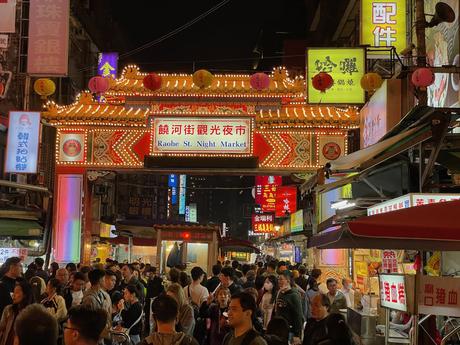 The illuminated gateway of Raohe Street Night Market, adorned with lanterns and decorative lights, with a lively crowd exploring food stalls below.