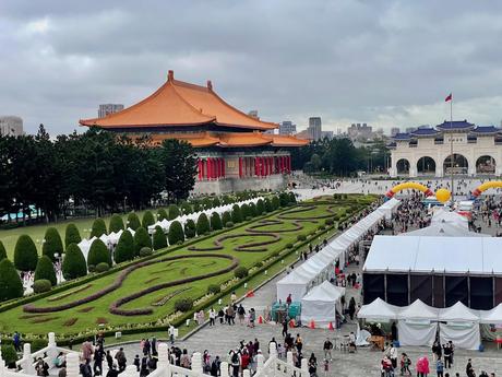 A view of the National Theater in Taipei with its orange-tiled roof and red columns, surrounded by neatly trimmed green hedges and flower gardens, and white event tents set up nearby.