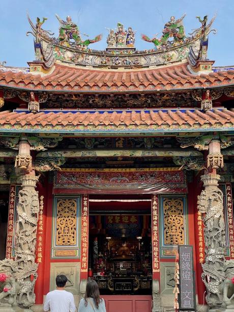 A grand temple façade featuring ornate details, colorful dragon sculptures on the roof, and red columns. A couple is admiring the elaborate entrance adorned with lanterns.