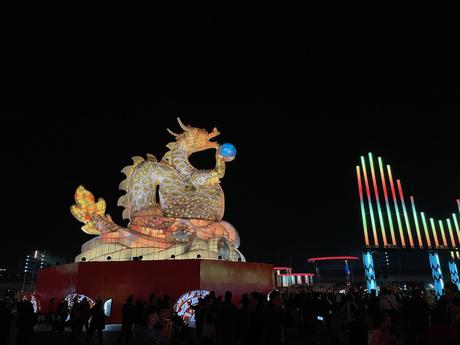 A large illuminated dragon sculpture at the Taiwan Lantern Festival holding a glowing sphere at a nighttime festival, with colorful light installations and a crowd of spectators nearby.