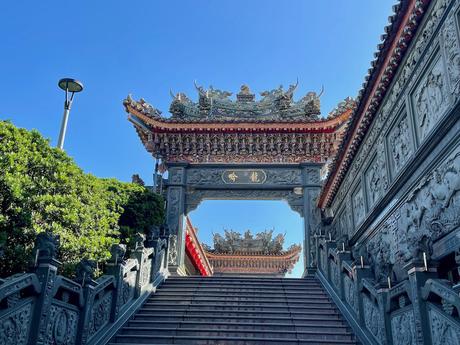 A grand temple gate with intricate carvings and Chinese characters, leading up a stone staircase under a clear blue sky.