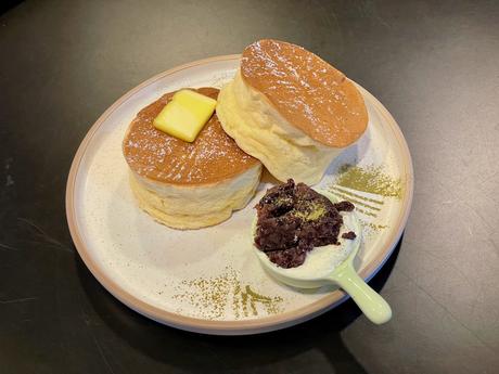 A plate of fluffy Japanese-style soufflé pancakes topped with a square of butter and served with a side of whipped cream and chocolate crumble.