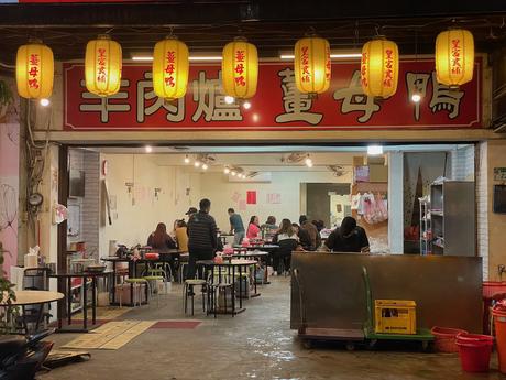 A warmly lit street-side restaurant entrance decorated with red and yellow hanging lanterns, with diners visible inside.