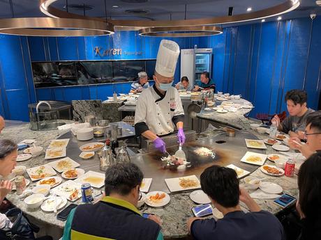 A chef wearing a tall hat and mask prepares food on a teppanyaki grill, surrounded by diners enjoying their meals at a circular table.