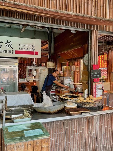 A local Taiwanese food vendor behind a counter, cooking and arranging buns and dumplings. The stall features bamboo decor and a menu in Chinese characters.