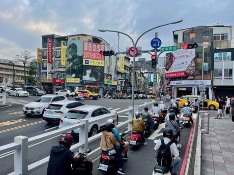 A busy street intersection in Taiwan with motorbikes at a stoplight, colorful billboards, and bustling urban scenery in the background.