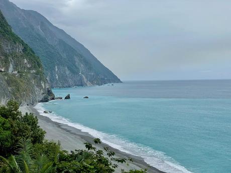 A peaceful shoreline with gray sand and vibrant blue water, framed by lush vegetation and a backdrop of misty mountains.