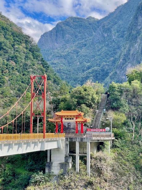 A detailed view of a red suspension bridge and the ornate pagoda entrance surrounded by greenery, with mountain peaks in the background.