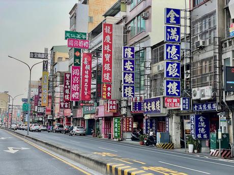 A typical Taiwanese city street lined with colorful storefront signs in Chinese characters advertising jewelry shops and wedding services. Cars and scooters fill the busy road.