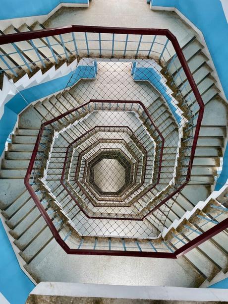 A spiraling staircase viewed from above, showcasing a geometric pattern of blue walls, red railings, and intricate symmetry.