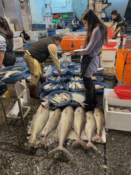 Vendors sorting large fish laid out on ice, with baskets of smaller fish nearby, and people working in the background in a vibrant seafood market.