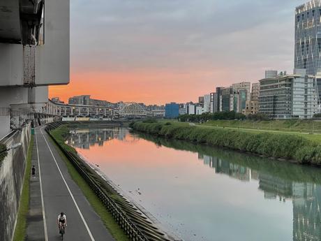 A riverside cycling and walking path in Taipei at sunset, with city buildings and a bridge reflected in the calm water, under a pink and orange sky.