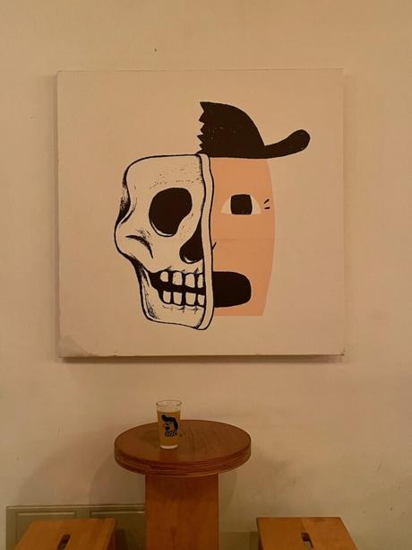 A piece of abstract art on a wall featuring a split face—one side a skull, the other a stylized human face wearing a hat—with a small glass on a wooden table below it.