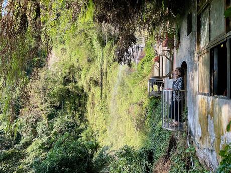 A woman standing on a small balcony of a stone temple built into a cliffside, surrounded by dense greenery and cascading water.