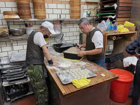 Two men working in a traditional kitchen preparing dumplings, with stacks of steamers and trays of dumplings on a wooden workbench covered in flour.