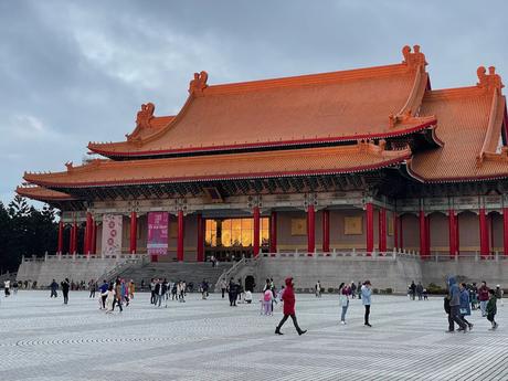 A grand public building with traditional Chinese architecture at dusk with its orange-tiled roof and striking red pillars, surrounded by an open plaza with scattered visitors.