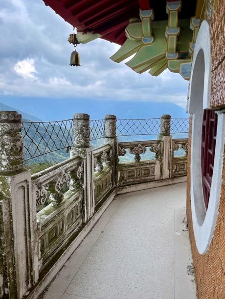 The upper balcony of Ci'en Pagoda, with traditional Chinese architecture, decorated with ornate stone carvings and a hanging bell, overlooking a mountainous landscape under a cloudy sky.