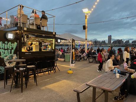 A riverside food and drink area at Pier 5 in Taipei with people dining at wooden tables, food stalls lit up under string lights, and city buildings visible across the water in the distance.