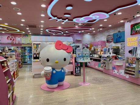 A brightly lit convenience store featuring a large Hello Kitty statue in a blue overall, holding a cup, with pink and pastel decor and shelves filled with products. Neon lights and signage add a playful touch.