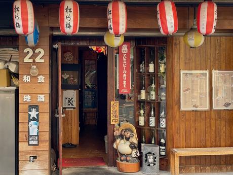 The entrance of a traditional-style izakaya restaurant with a wooden facade, Japanese lanterns, a raccoon dog statue, and shelves of sake bottles displayed by the door.