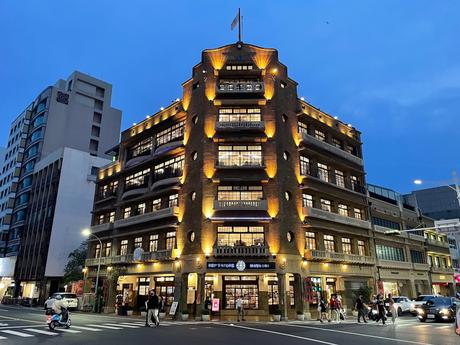 The iconic Hayashi Department Store in Tainan, illuminated at dusk, with warm yellow lighting, arched windows, and a lively street scene with pedestrians and vehicles.